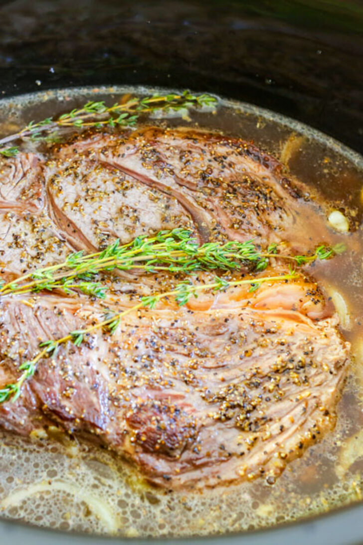 a photo of a chuck roast in the crockpot with broth and herbs