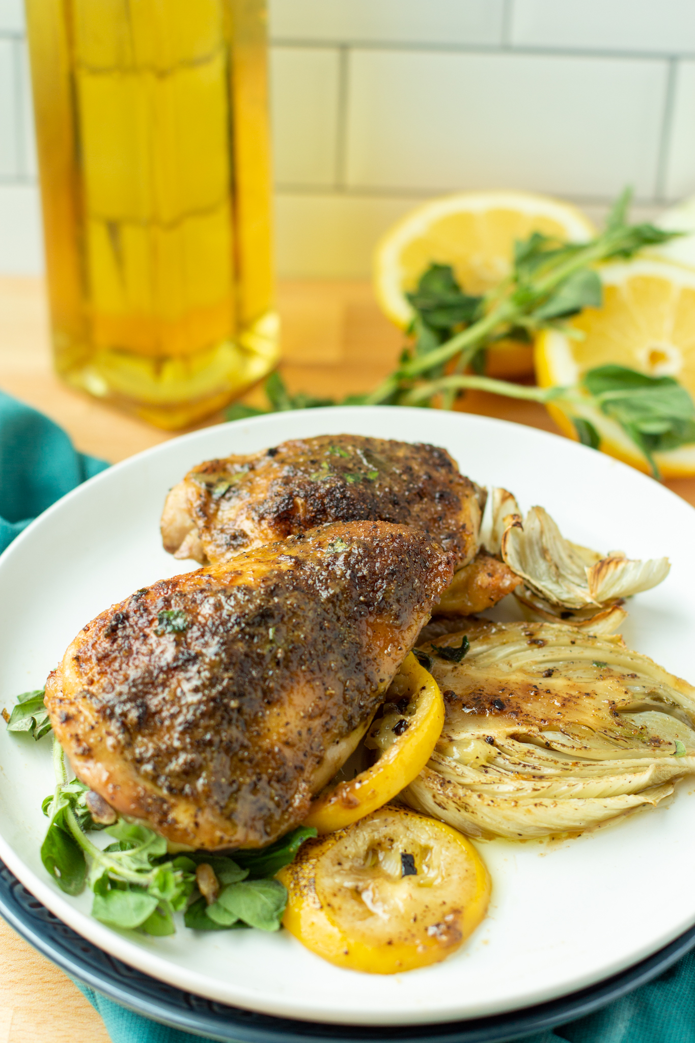 A photo of roasted chicken thighs on a plate with roasted fennel, lemon and olive oil in the background