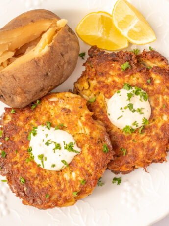 an overhead photo of crab cakes topped with aioli and fresh herbs, with lemon wedges and a baked potato