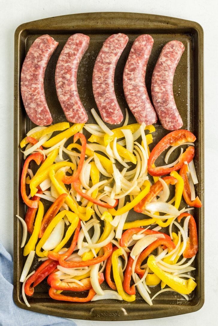 an overhead photo of raw bratuwrst, peppers and onions on a baking sheet