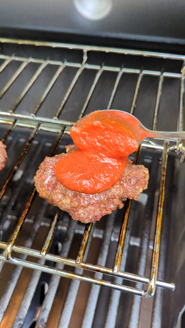 a photo of a burger on the grill being topped with a spoonful of pizza sauce