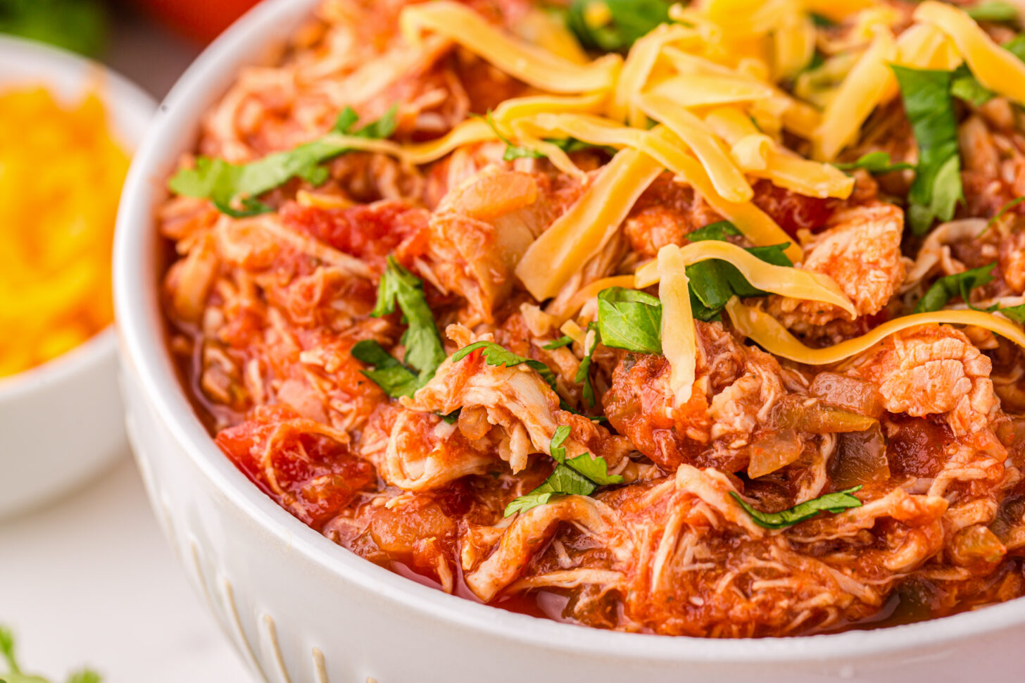 A close-up of a white bowl filled with shredded chicken in a tomato-based sauce, topped with shredded cheese and chopped fresh herbs. The dish looks savory and flavorful, with additional cheese visible in a smaller bowl in the background.