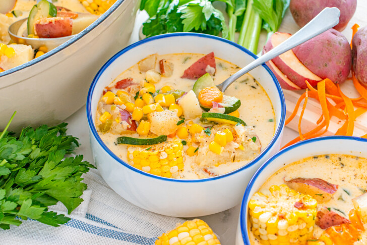 a bowl of corn chowder surrounded by herbs and corn cobs